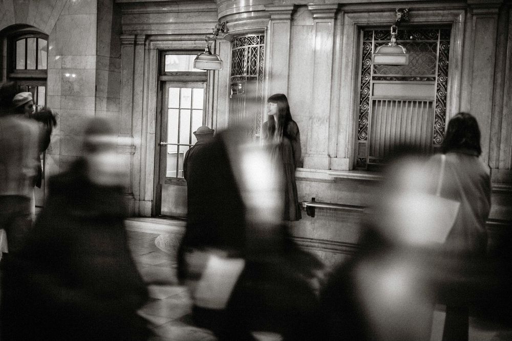Black and white photo of blurred pedestrians walking past a woman standing still in a grand building interior.