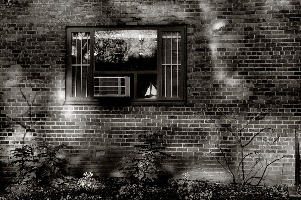 Black and white photo of a brick wall with a window reflecting trees, an AC unit inside, and small sailboat figurine visible.