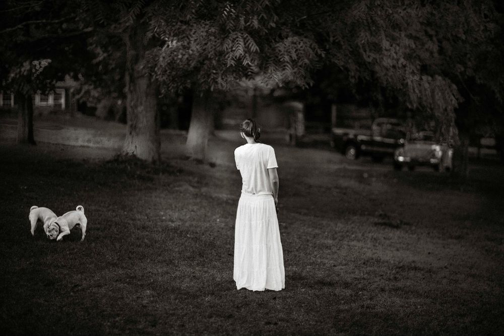 Woman in white dress standing in park with small dog, vintage car in background, black and white photo.