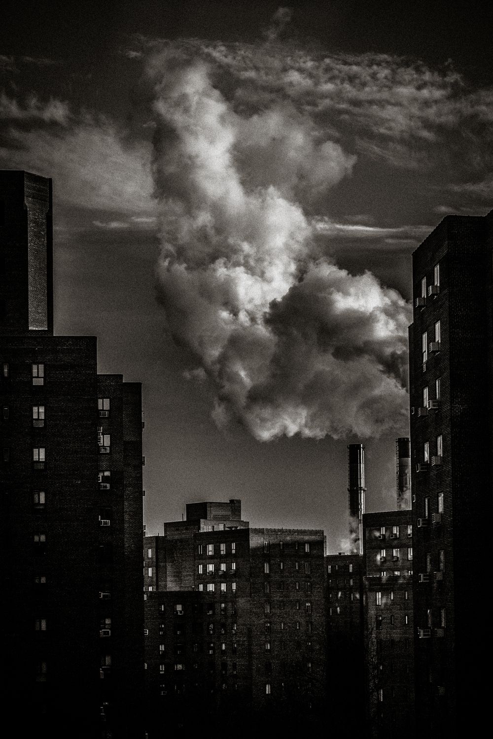 Black and white cityscape with dramatic smoke billowing from a chimney between dark urban buildings.