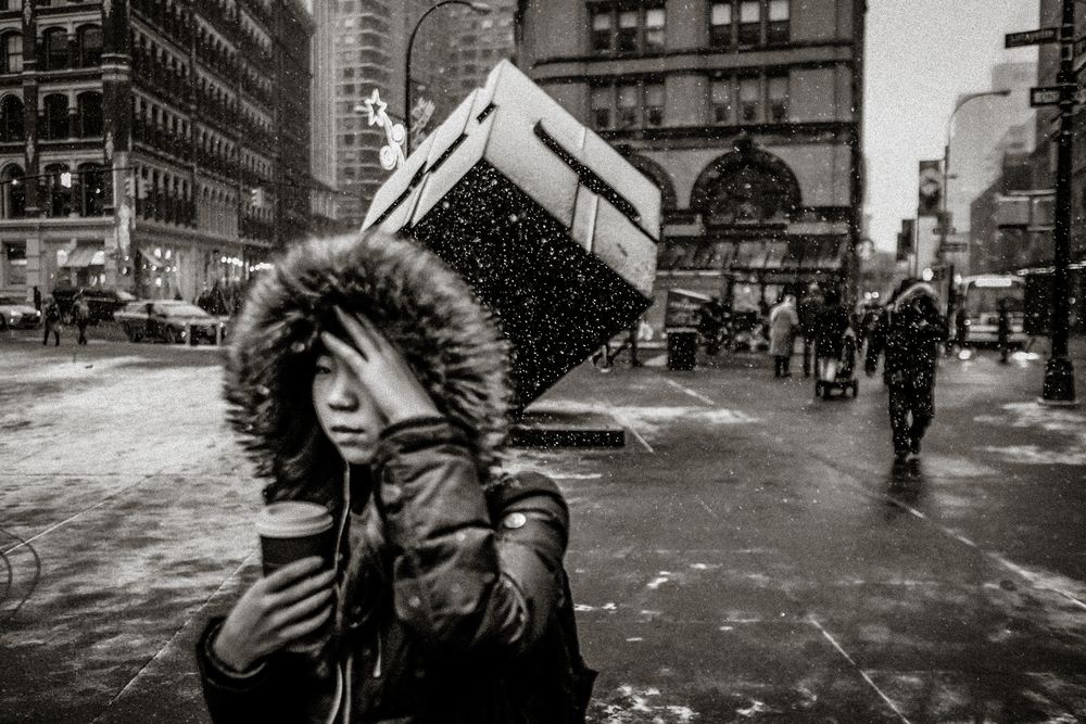 Black and white street photo of a woman in a fur-hooded coat holding coffee in snowy New York City.