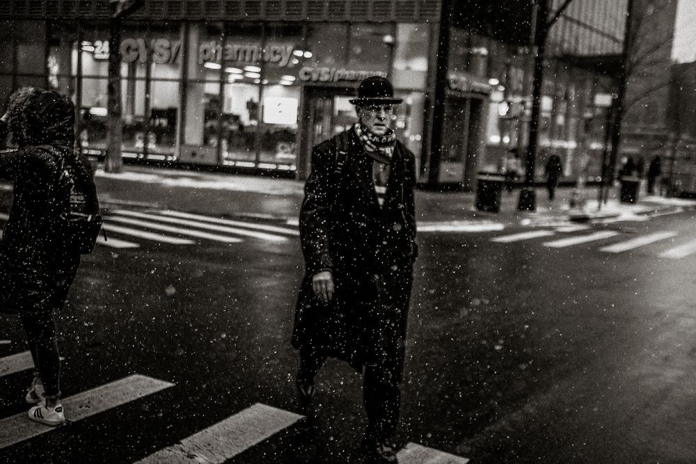 Elderly man in black coat and hat crossing a snowy street at night near a CVS pharmacy.