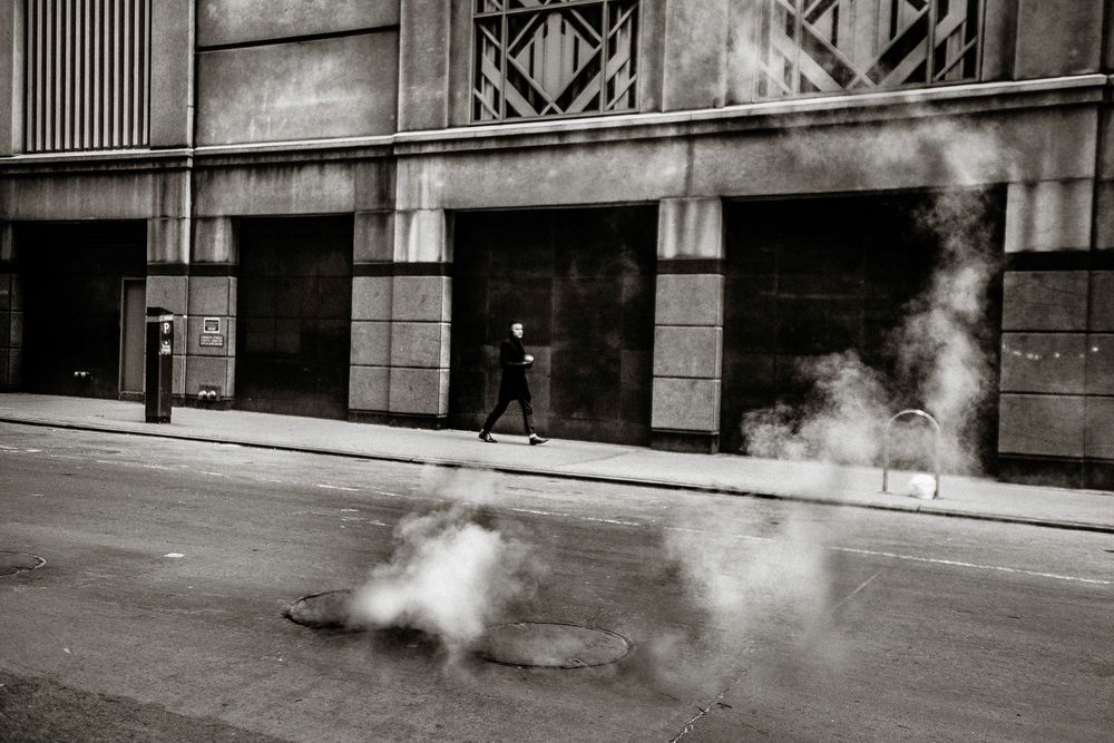Black and white photo of a person walking past a building with steam rising from a manhole cover on a city street.
