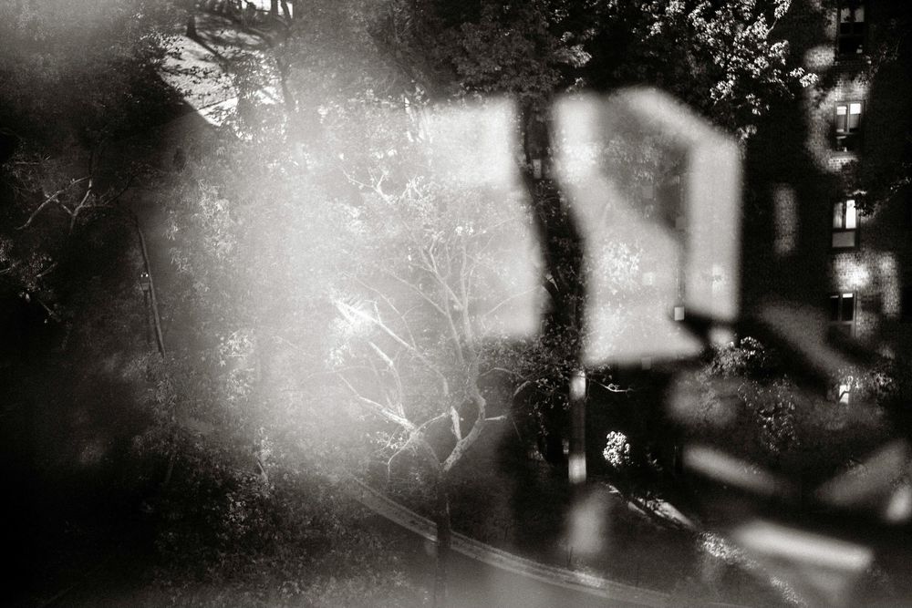 Ethereal black and white photo of trees and buildings with dramatic light flare creating a ghostly, dreamlike effect.