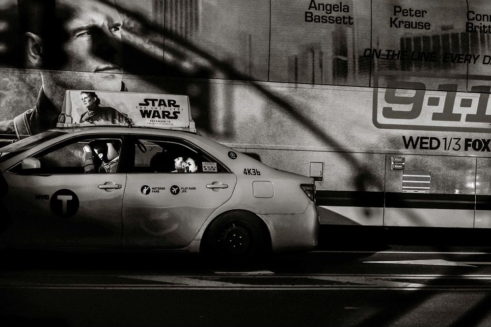 Black and white photo of a NYC taxi cab with a Star Wars advertisement, parked beside a bus on a city street.