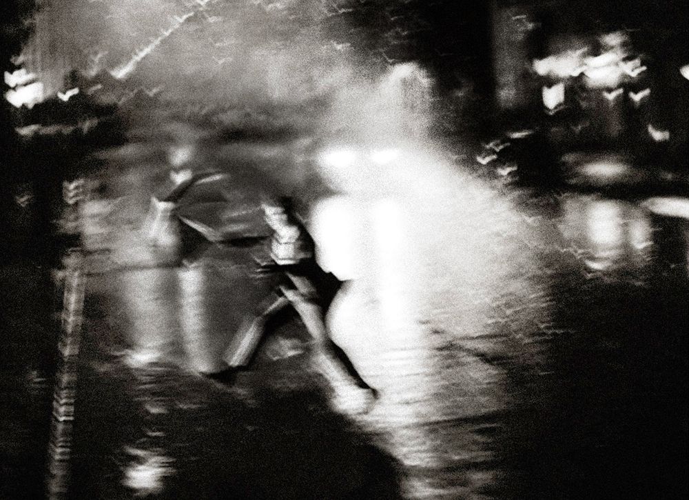 Blurred black and white photo of a person running through rain-soaked streets at night with glowing lights.