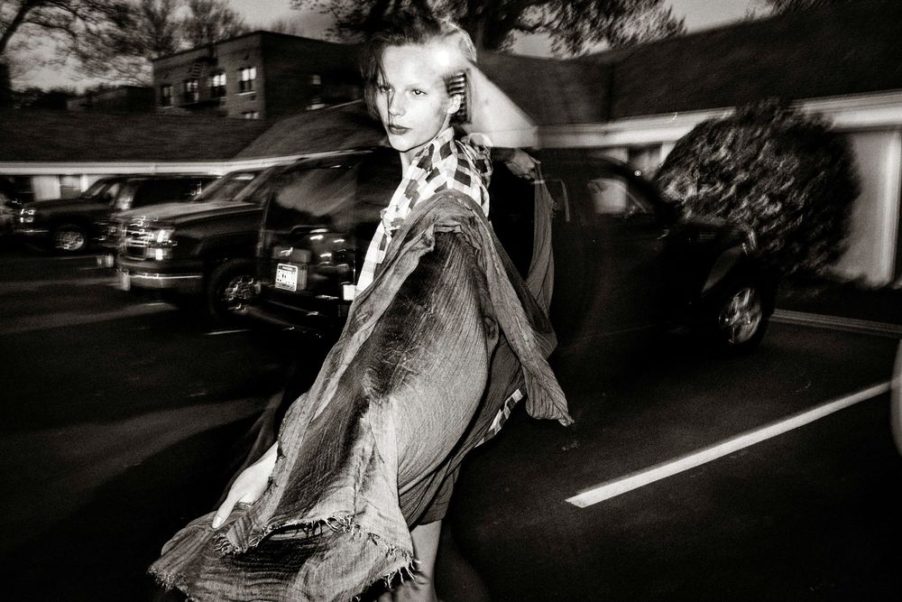 Black and white fashion photo of a blonde woman in a flowing coat walking through a busy street at night.