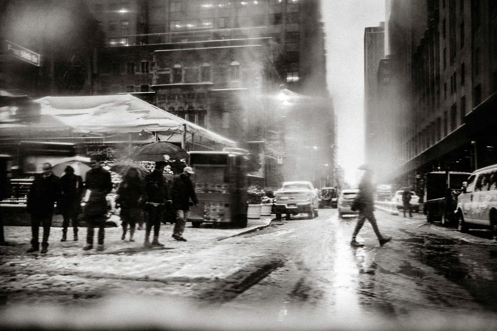 Black and white photo of snowy New York City street with pedestrians and cars amid steam and glowing lights.