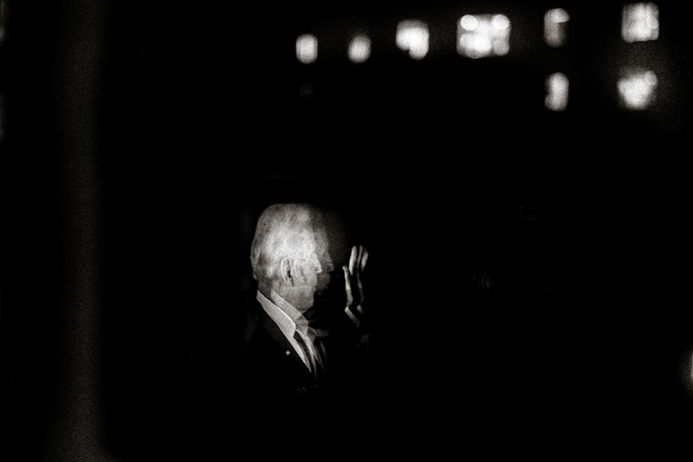 Dramatic black and white portrait of an elderly man in a suit, illuminated against a dark background.
