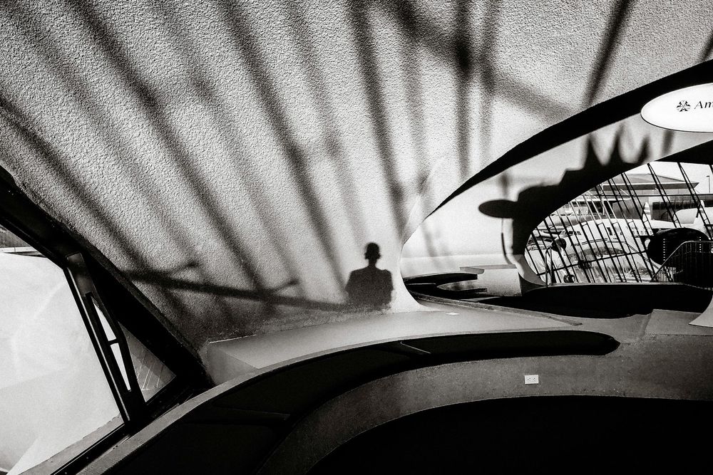 Black and white photo of a shadow silhouette on a car hood with dramatic light streaks in an urban setting.