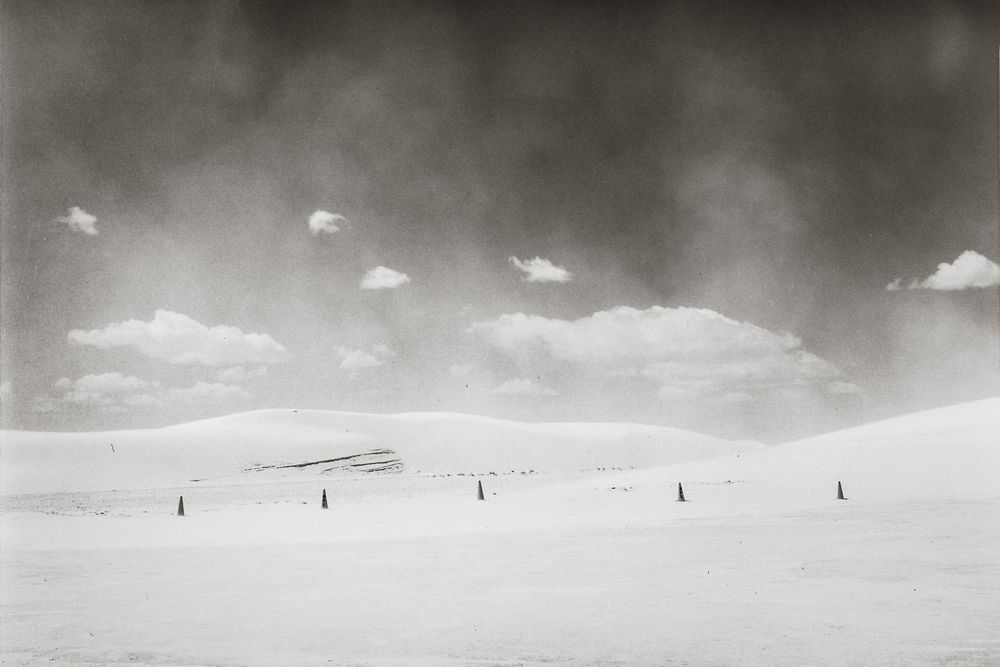 Black and white photo of vast snowy dunes under dramatic cloudy sky with small markers dotting the landscape.