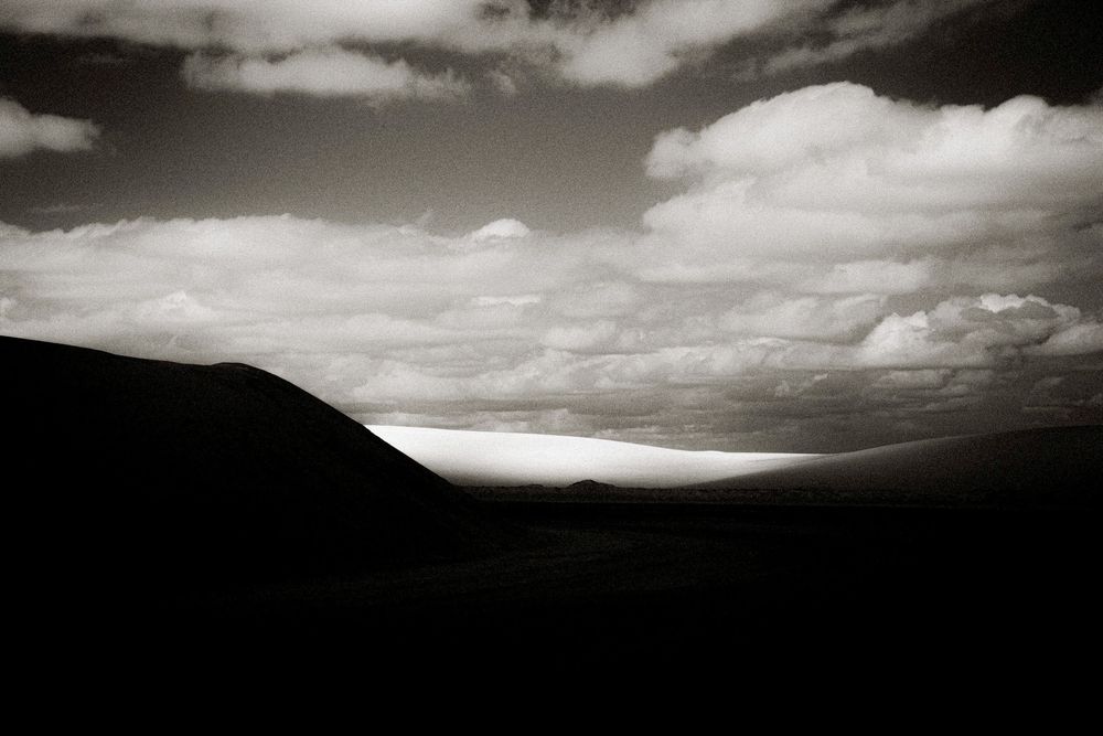 Black and white landscape photo of dark rolling hills under dramatic cloudy sky with light breaking through on horizon.