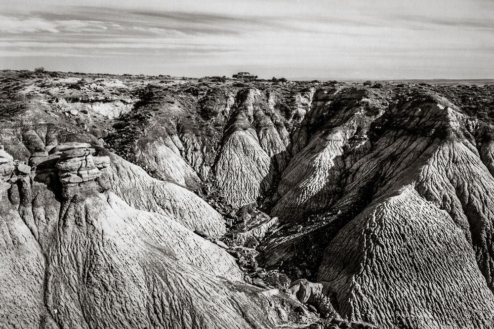 Black and white aerial view of eroded badlands rock formations with dramatic ridges and valleys under a cloudy sky.