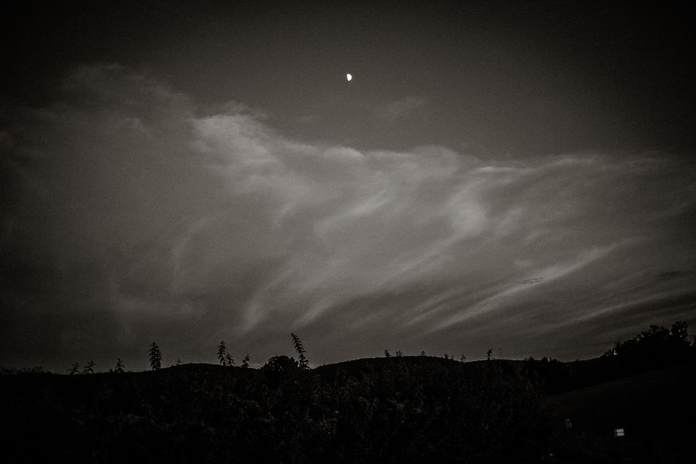 Dark moody night sky with wispy clouds and a crescent moon above a silhouetted hillside landscape.