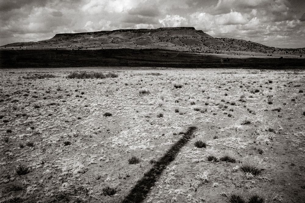 Black and white desert landscape with mesa, sparse scrubland, and dramatic shadow leading toward distant mountain under cloudy sky.