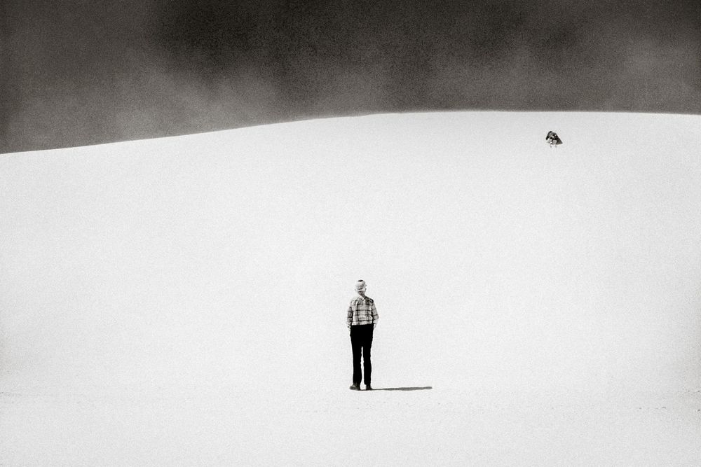 A lone person stands on a vast snowy white sand dune in a minimalist black and white landscape photo.