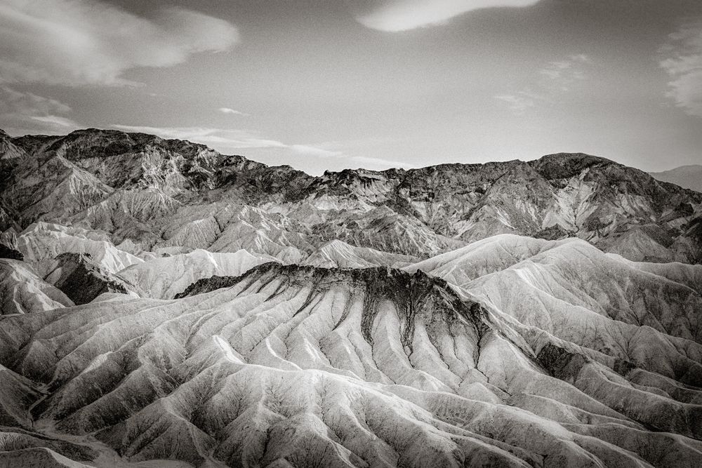 Black and white aerial view of dramatic eroded badlands terrain with layered ridges and valleys under cloudy sky.