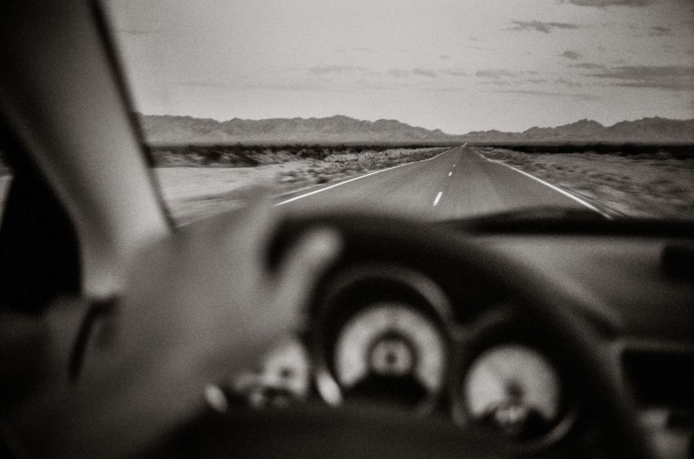 Black and white view from inside a car driving on a long straight desert highway toward distant mountains.