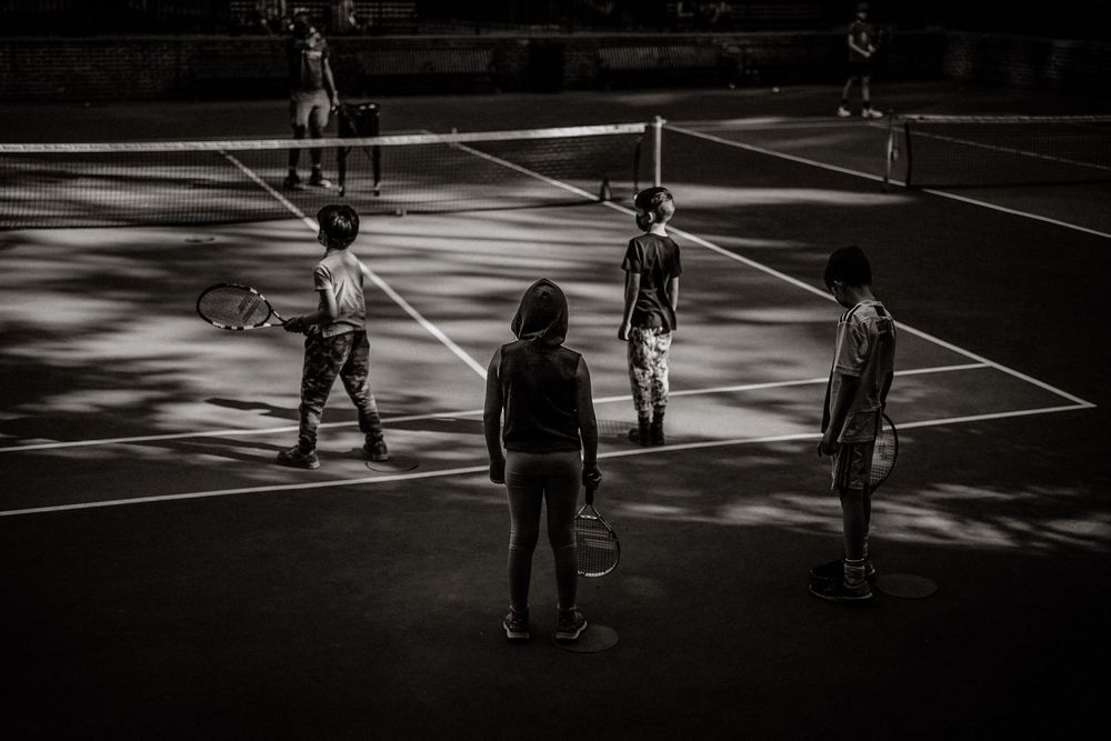 Black and white photo of children playing tennis on an outdoor court, holding rackets during a lesson.