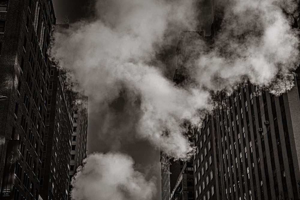 Black and white photo of billowing steam rising between towering city skyscrapers in an urban canyon.