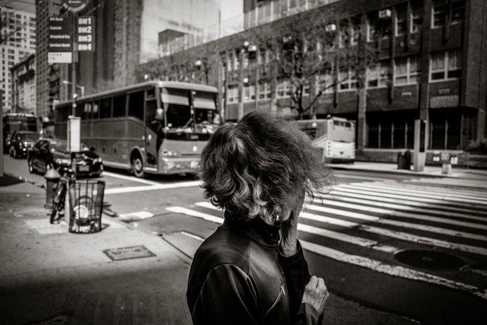 Black and white street photo of a woman with windswept hair on a busy urban intersection with buses and buildings.