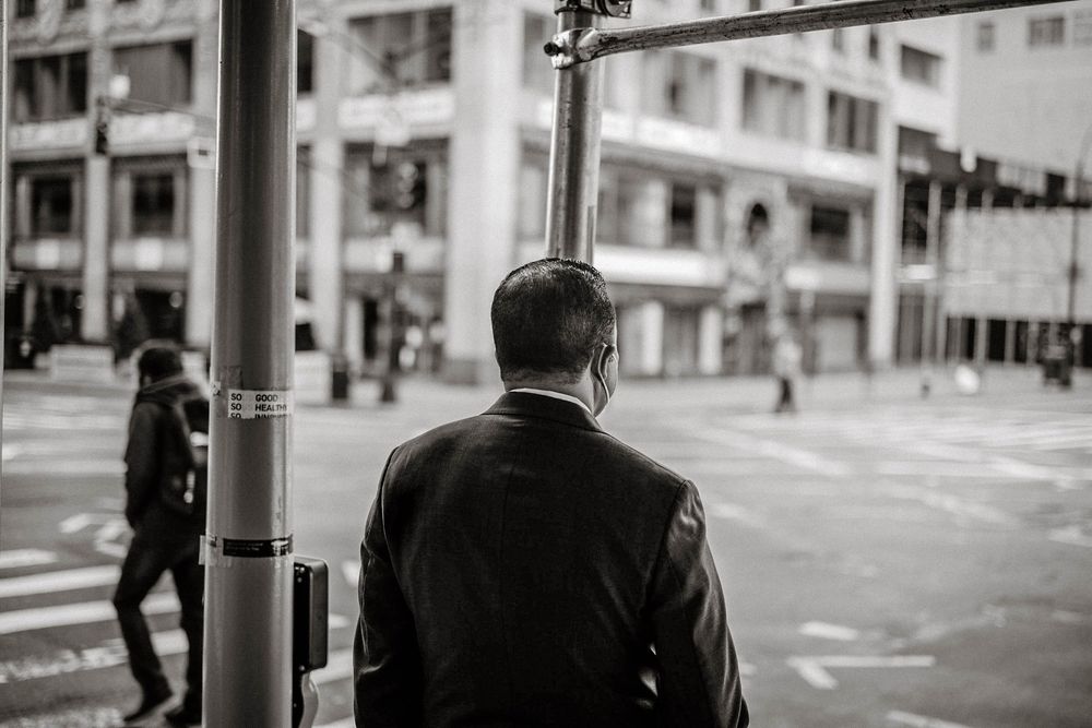 Black and white photo of a man in a leather jacket waiting at a city street corner with urban buildings.