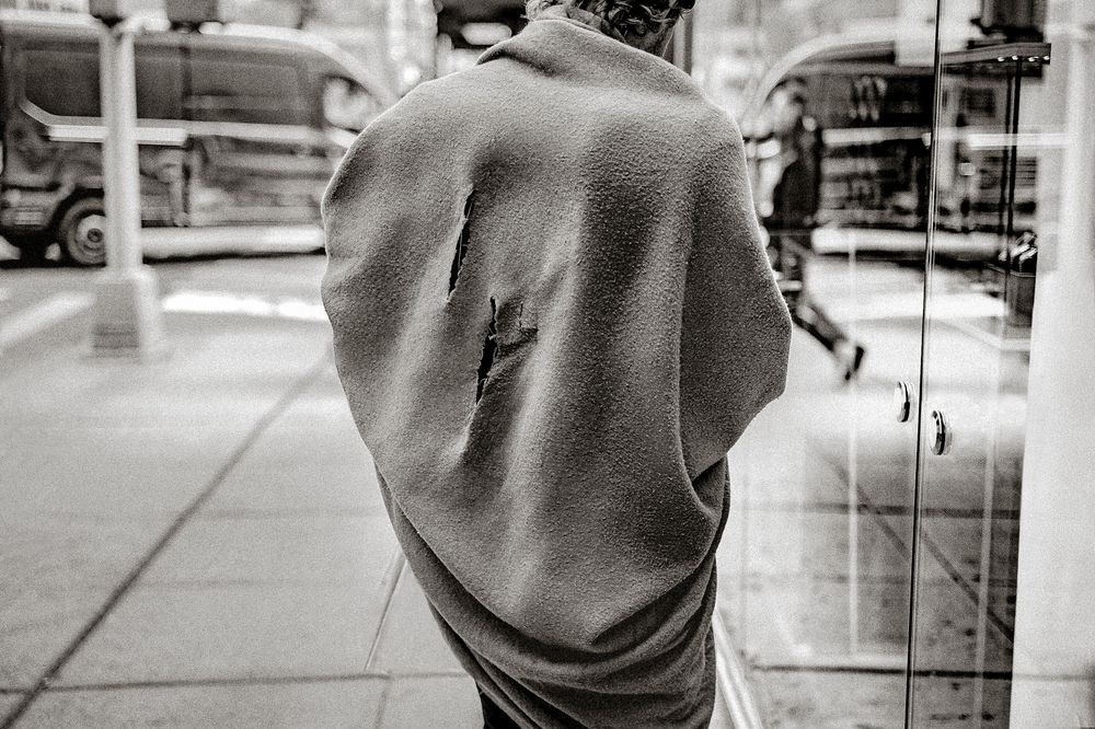 Black and white photo of person wearing gray hoodie with lightning bolt design on back, standing on city sidewalk.