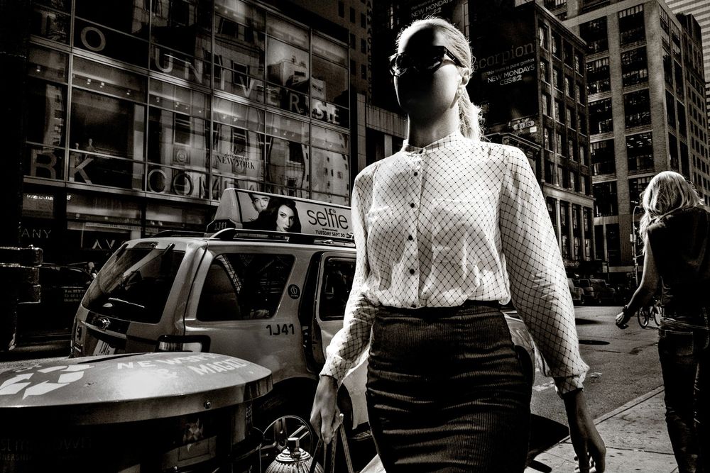 Black and white street photo of a woman in a white blouse and skirt walking confidently in a busy city.