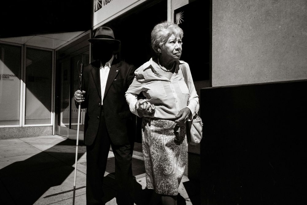 Black and white photo of an elderly couple walking together outside a building, man holding a cane.