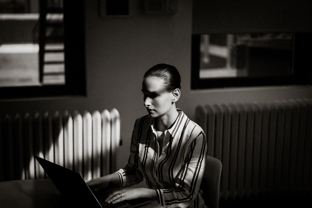 Black and white photo of a focused woman working on a laptop in a dimly lit room with a radiator.
