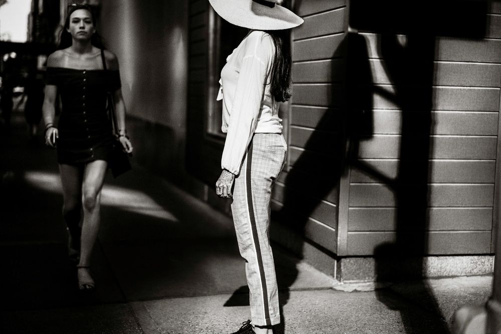 Black and white street photo of a woman in a wide-brim hat and striped pants walking near a building.