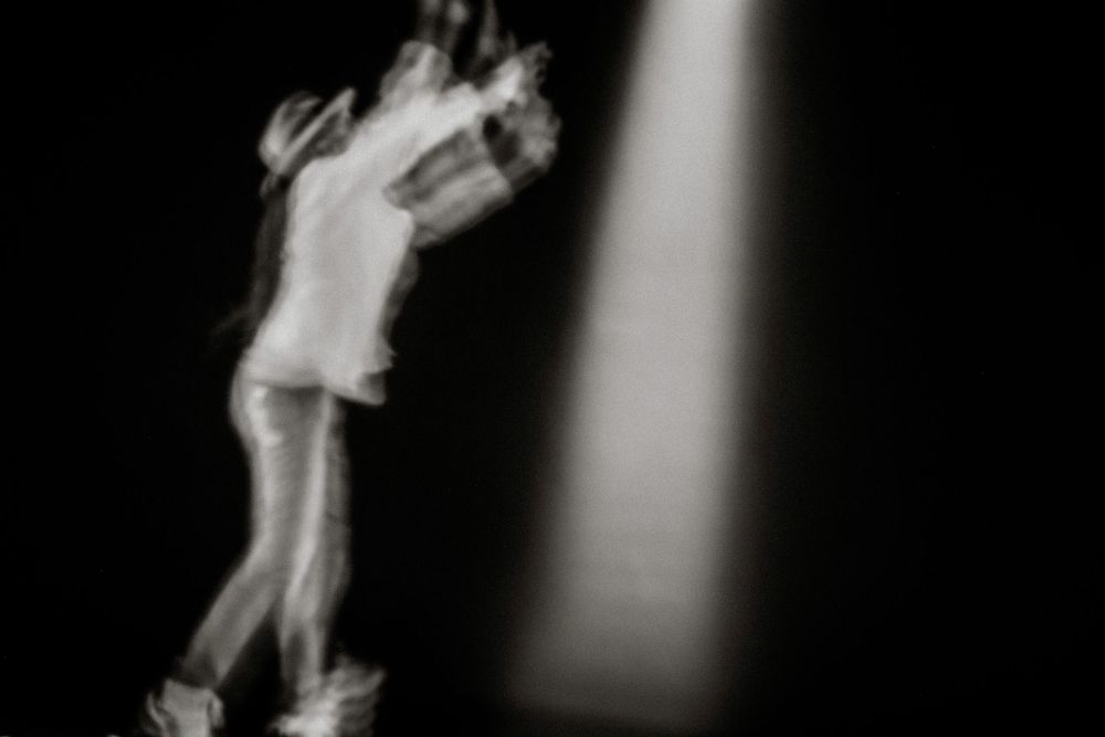 Blurred black and white photo of a performer dancing under a spotlight on a dark stage.