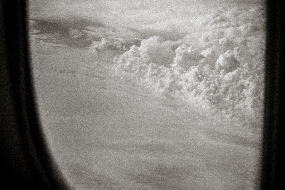 Black and white aerial view through airplane window showing dramatic cloud formations above the earth.