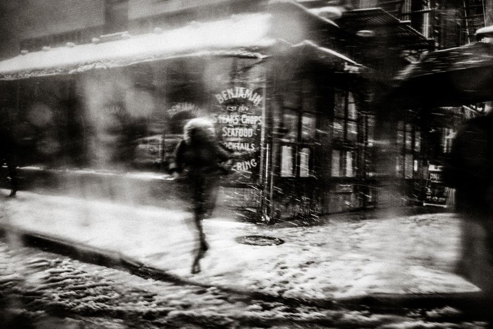 Blurry black and white photo of a person walking past a restaurant in a snowy urban street at night.