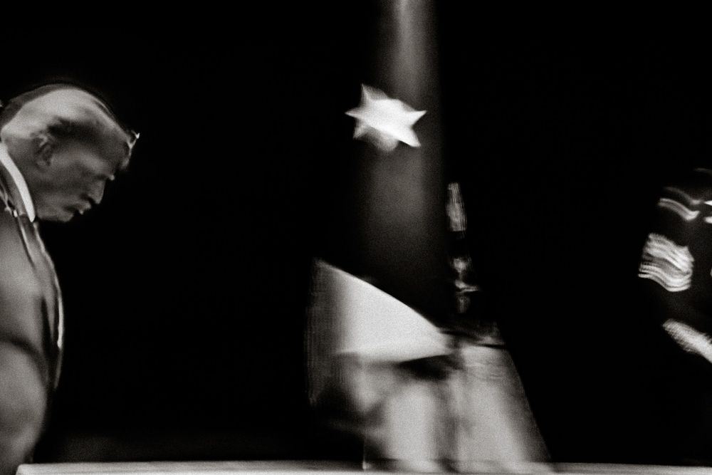 Black and white photo of a man in suit bowing head near a blurred American flag in dramatic lighting.