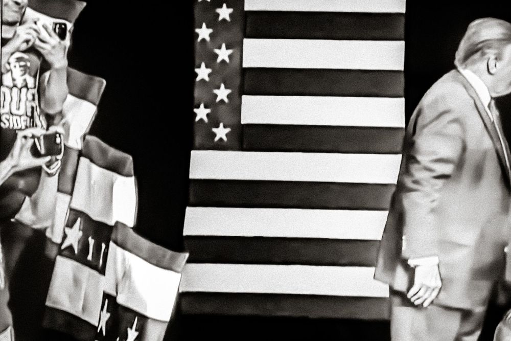 Black and white photo of a man standing before a large American flag at a political rally event.