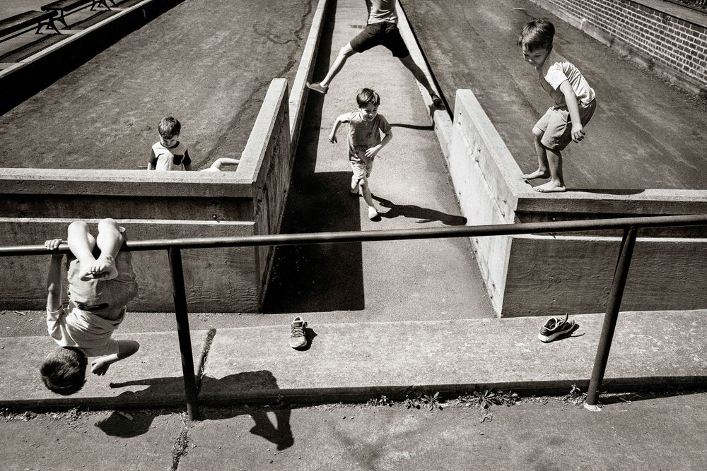 Black and white photo of children playing on a concrete skate park ramp, captured from an elevated angle.