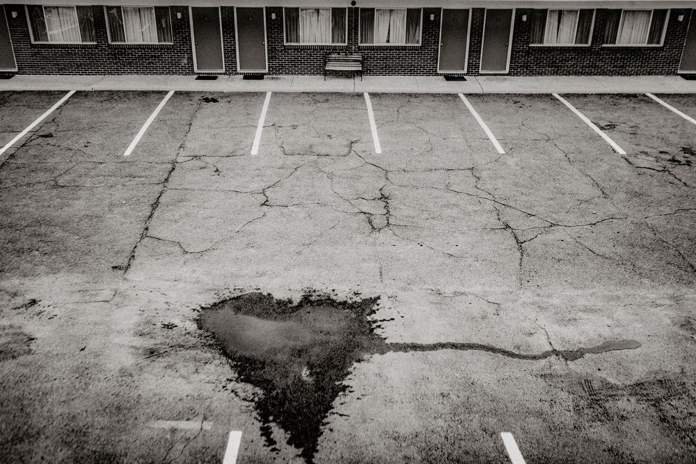 Black and white photo of an empty cracked parking lot with a puddle near a motel building.