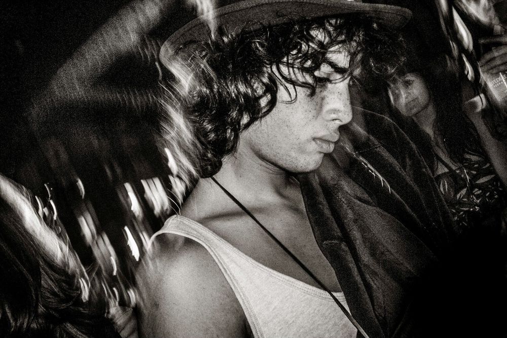 Black and white photo of a young man with curly hair wearing a tank top and necklace at a crowded event.