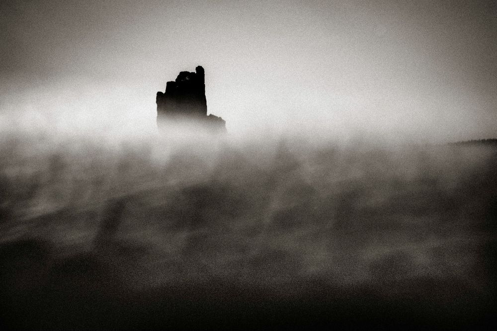 A dark silhouette of ancient ruins emerges through swirling fog in a dramatic black and white landscape.