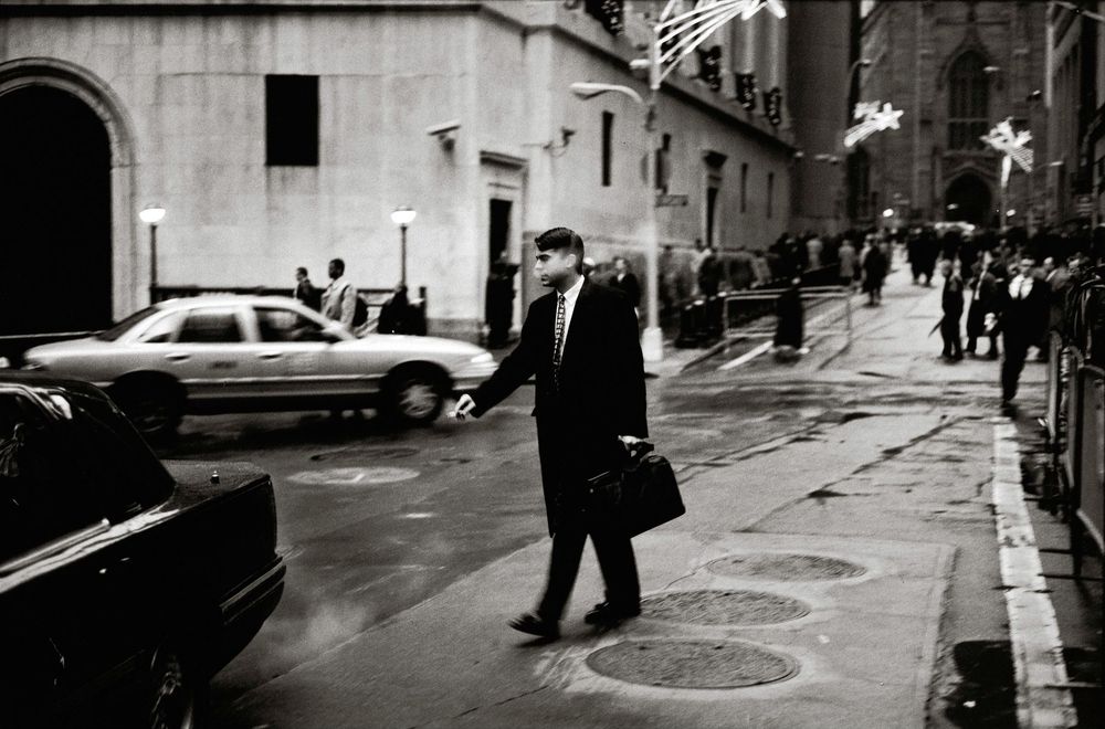 Black and white photo of a businessman carrying a briefcase crossing a wet city street on a rainy day.