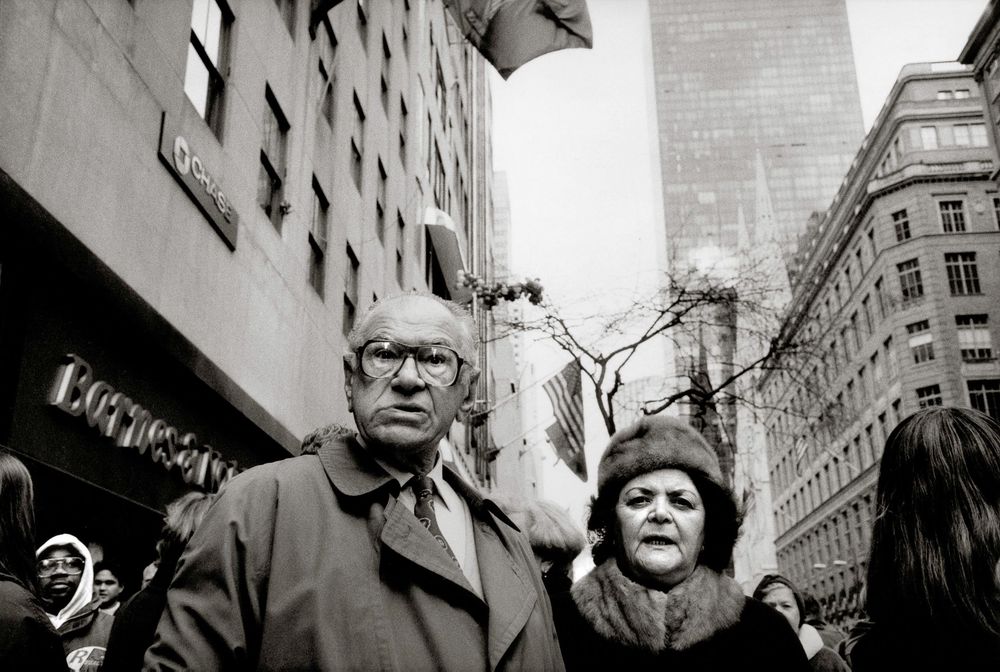 Black and white photo of an elderly couple standing on a busy urban street, with tall city buildings visible in the background.