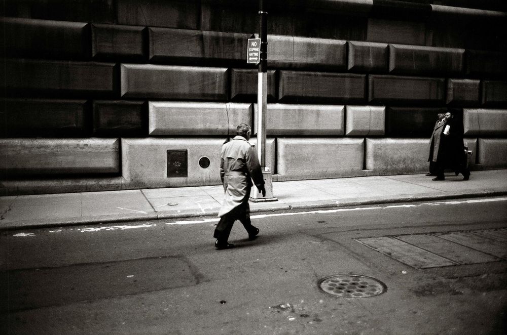 Black and white street photo of a person walking past a large stone building on an urban sidewalk.