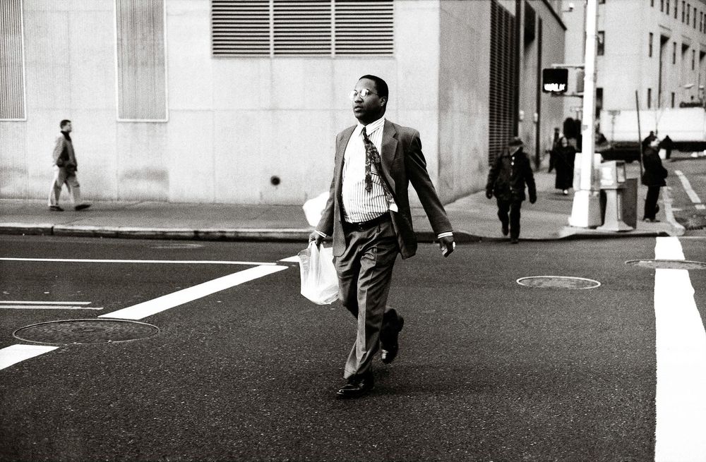 A man in a suit crosses a city street carrying a bag in a black and white urban street photography shot.