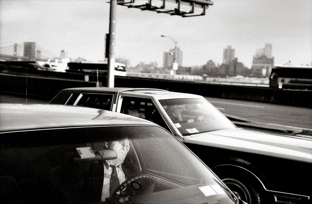 Black and white photo of an elderly man driving a classic car on a busy highway with a city skyline in the background.