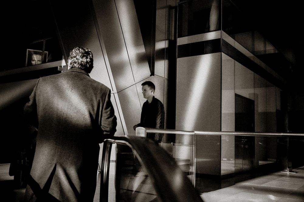 Two men in a dramatic black and white scene on a staircase, one in a suit facing a glass building at night.