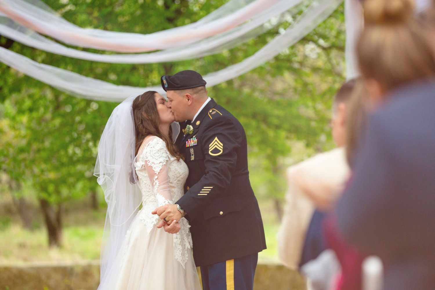 Military groom in Army dress uniform kisses bride in white lace gown and veil at outdoor wedding ceremony.