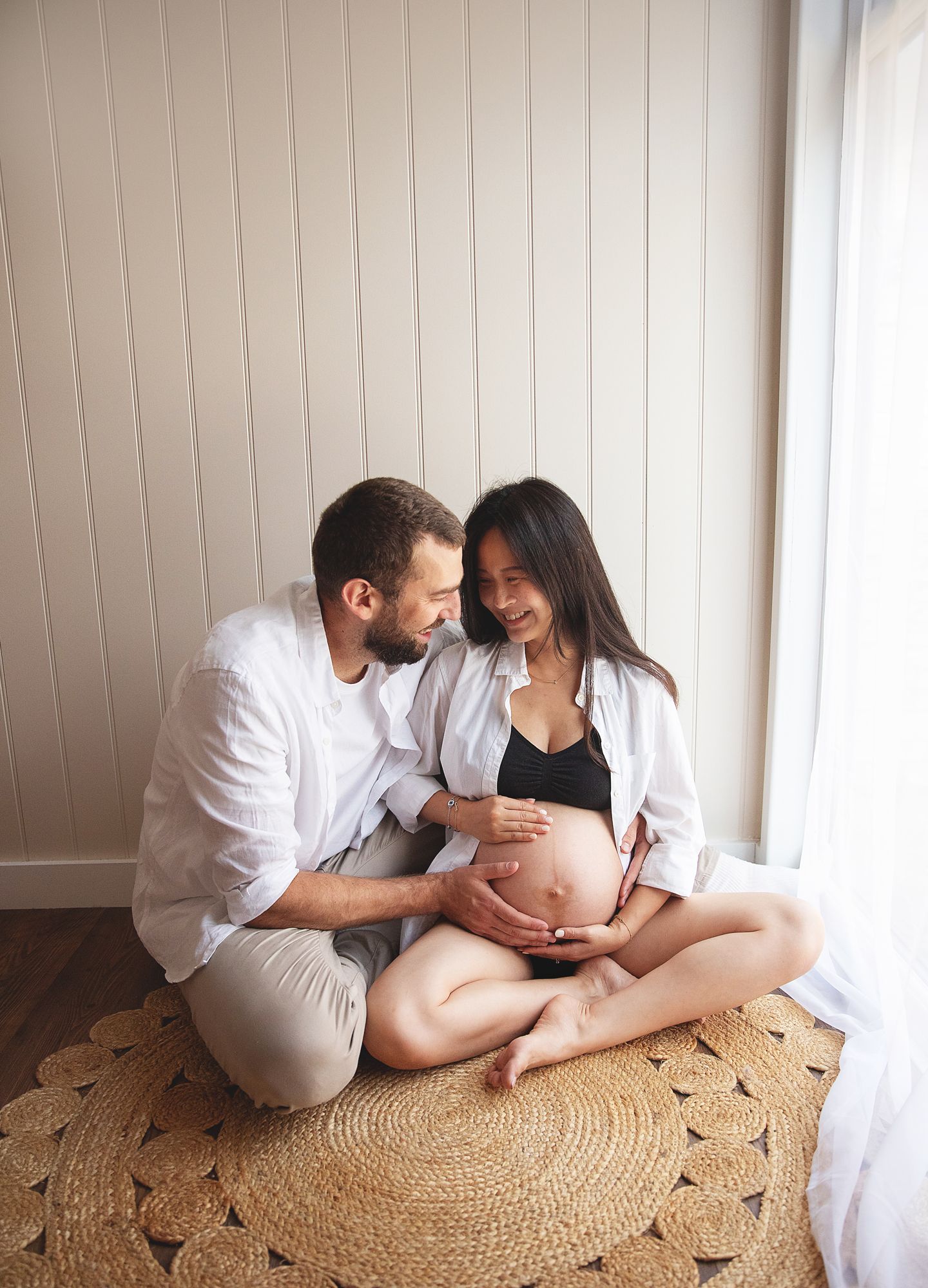Happy couple sitting on a rug, woman with pregnant belly, man in white shirt. During a maternity photoshoot in South East London