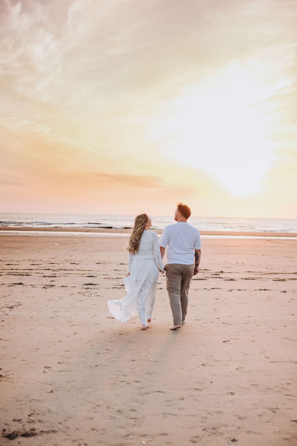 Couple holding hands walking on beach at sunset during a maternity photoshoot, woman in white dress and man in white shirt with gray pants.