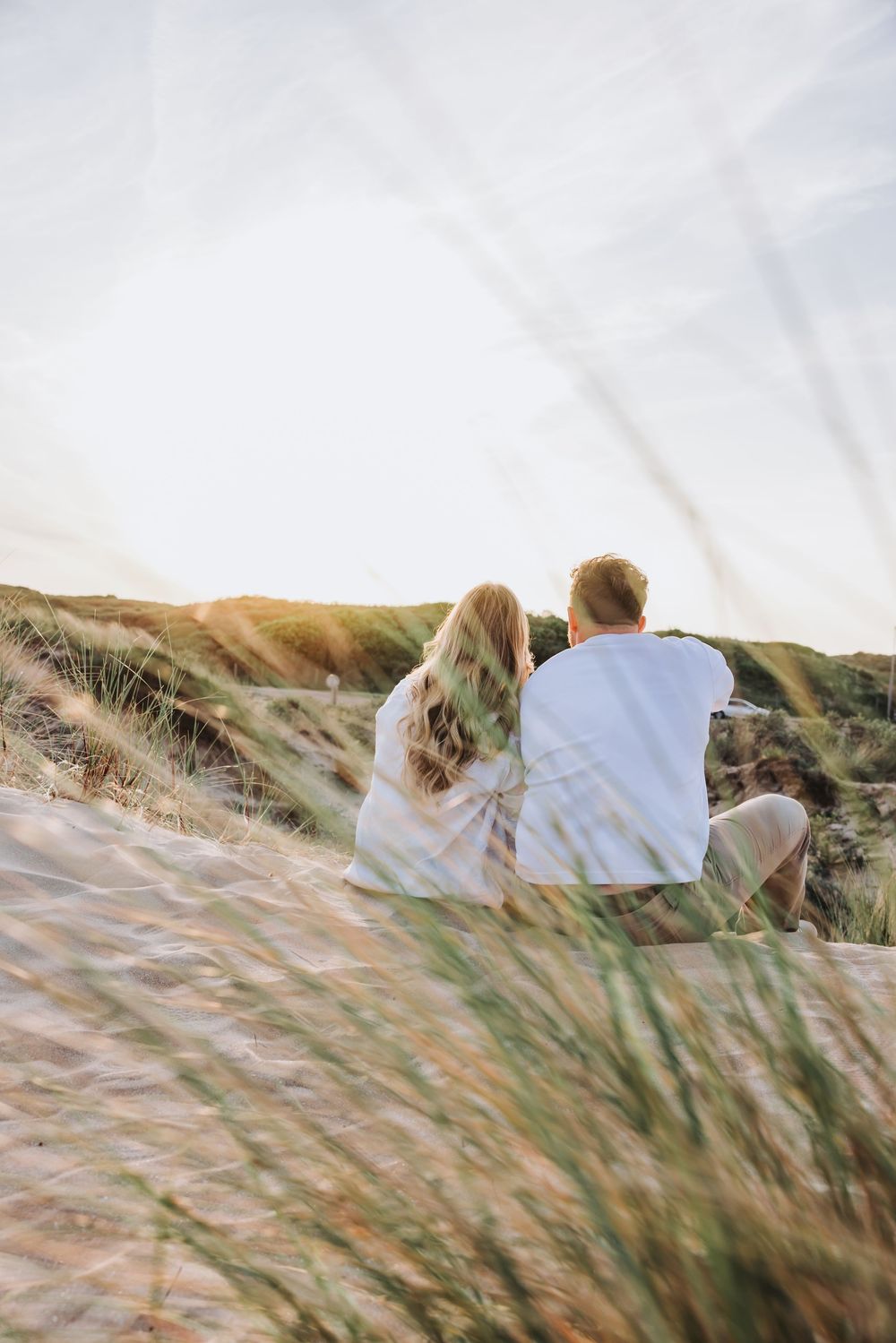 Couple sitting together on sandy dunes with beach grass, viewed from behind at golden hour sunset.